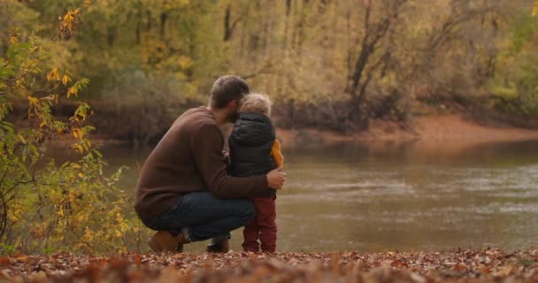 Pai caucasiano e filho pequeno estão descansando na margem da lagoa da floresta no outono, desfrutando da natureza e do clima quente no outono. — Vídeo de Stock