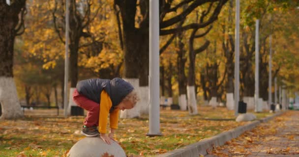 Herbsttag im Park, niedliches kleines Kind spielt, lächelt und zeigt der Kamera die Zunge, tagsüber in voller Länge aufgenommen — Stockvideo