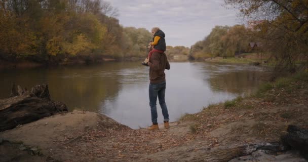 Vader laat zijn zoontje natuur en herfstlandschap zien, kind op de schouders houden, aan de kust van het meer staan — Stockvideo