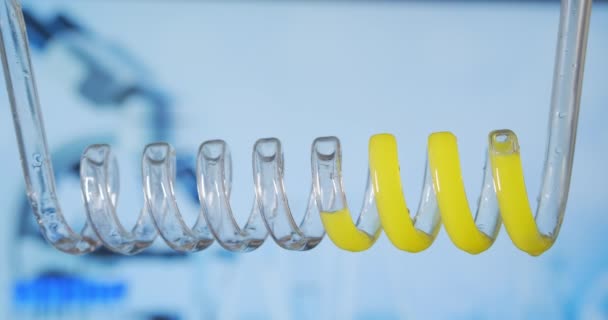 Nahaufnahme und Zeitlupe einer gelben Flüssigkeit, die sich horizontal von rechts nach links in einem wissenschaftlichen Glaskondensator spiralförmig bewegt. Entwicklung und Suche nach einem Impfstoff. Chemische Forschung am Coronavirus. Drogentests. — Stockvideo