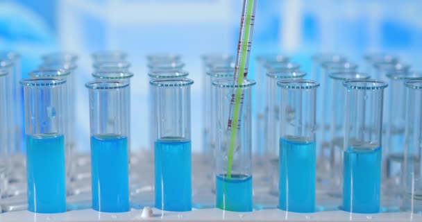 Вчений вводить хімічні речовини в пробірки. Випробувальна трубка кольорової рідини на лабораторному столі на фоні скляних колб. наливається зі скляної піпетки в пробірку, освітлену синім світлом — стокове відео