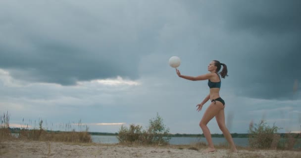 スレンダーなビーチバレー選手の女性は砂のコートでボールを提供しています,曇りの空に対する完全な長さショット — ストック動画