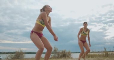 Plaj voleybol takımındaki iki kadın bulutlu yaz gününde sahada antrenman yapıyor, sporcular spor bikini giyiyor.