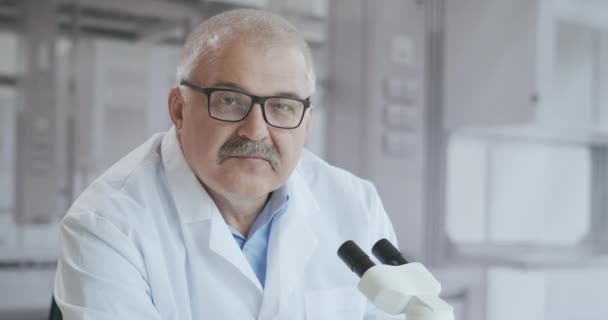 Portret een witte harige man lab arts met een microscoop kijkt serieus naar de camera en glimlacht dan in slow motion — Stockvideo