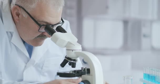 Pesquisador masculino caucasiano barbudo usando óculos de proteção e trabalhando com um microscópio spbas. cientista usando microscópio em um laboratório. Procurar vacina contra o coronavírus — Vídeo de Stock
