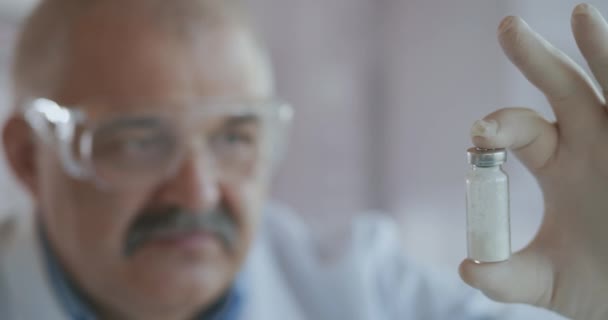Extrémní detailní záběr vědce v modrém respirátoru a ochranných brýlích, který vyvinul koronavirovou vakcínu obsahující ampulku bílého prášku. Narkotika a léky proti bolesti — Stock video