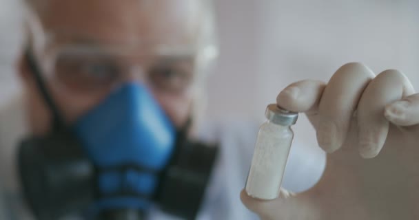 Ekstremalne zbliżenie naukowca w niebieskim respiratorze i okularach ochronnych, który opracował szczepionkę przeciw koronawirusowi zawierającą ampułkę białego proszku. Środki odurzające i przeciwbólowe — Wideo stockowe