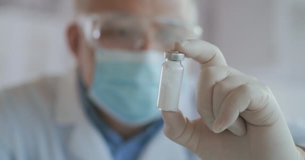 Close-up um cientista mascarado que desenvolveu uma vacina contra o coronavírus detém uma ampola de pó branco e examina a vacina. O médico olha para os antibióticos antes de usar — Vídeo de Stock