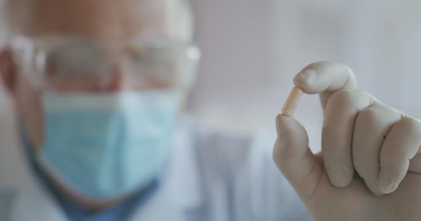 Extremo primer plano de un científico enmascarado desarrollando una vacuna contra el coronavirus sosteniendo una píldora blanca. El médico examina los analgésicos antivirales. Vitaminas. Nuevo tipo de medicamento — Vídeo de stock