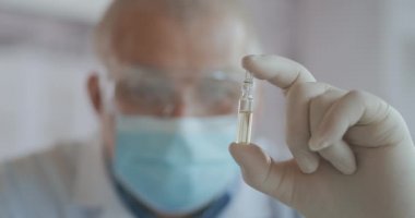 Koronavirüs aşısı geliştiren maskeli bir bilim adamı beyaz sıvı bir ampul tutuyor ve aşıyı inceliyor. Doktor uygulamadan önce ilaca bakar.