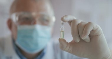 Coronavirus aşısının geliştiricisinin elinde bir ampul beyaz sıvı var. Aşıyı eldiven içinde inceliyor ve tutuyor.