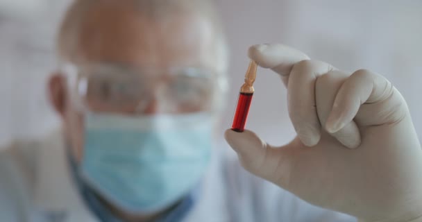 Close-up van de ontwikkelaar van het coronavirusvaccin met een ampul rode vloeistof die het vaccin onderzoekt en met handschoenen vasthoudt — Stockvideo