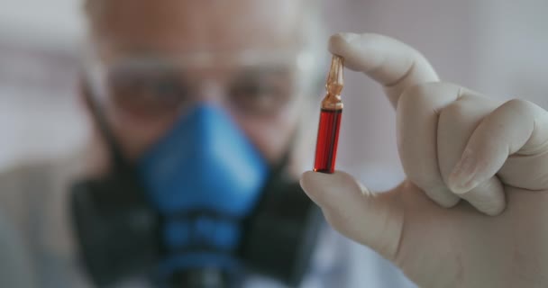 Extreme Close-up of a scientist in a blue respirator and protective glasses, a developer of a coronavirus vaccine holding a red ampoule. The doctor looks at the new vaccine. A new type of virus — Stock Video