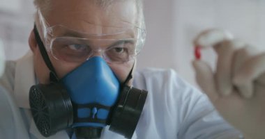 Bir ilaç şirketinde çalışan bir bilim adamı mavi solunum cihazı ve güvenlik gözlüğü takan bir ilaç geliştiricisi elinde yeni bir ilaç tutuyor kırmızı bir hap ve ona bakıyor.