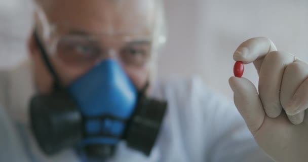 Um cientista de uma empresa farmacêutica um desenvolvedor de drogas usando um respirador azul e óculos de segurança segura um novo medicamento uma pílula vermelha e olha para ele — Vídeo de Stock
