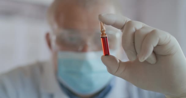 Un scientifique masculin tient une ampoule en verre rouge avec un vaccin et la regarde en gros plan au ralenti. Vaccinations — Video