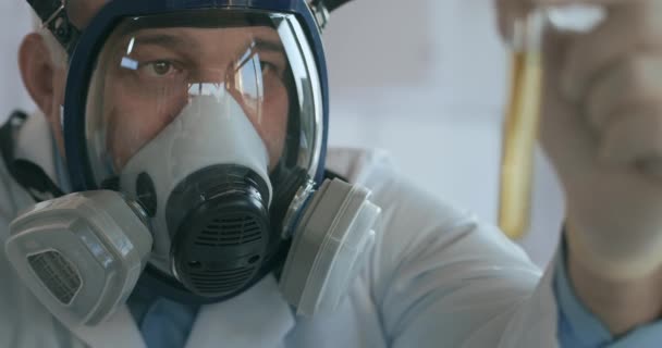 Close-up in het laboratorium een man in een glazen masker scherm houdt een glazen buis met een gele vloeistof tegen het virus en kijkt ernaar — Stockvideo