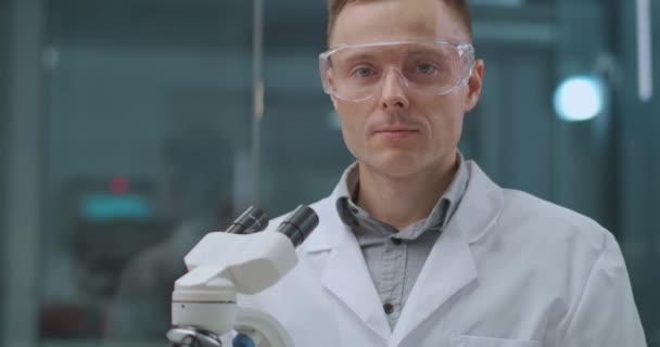 Портрет уверенного лаборанта-техника в рабочей комнате, в защитных очках, стоящего рядом с микроскопом — стоковое видео
