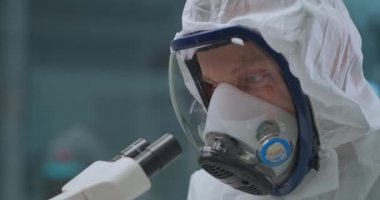 Mikrobiyolog, laboratuvarda mikroskopla tehlikeli bakterileri araştırıyor, kendini genel olarak koruyor ve solunum cihazıyla maske takıyor.