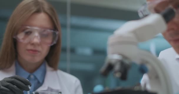 Equipo de jóvenes científicos está trabajando en laboratorio farmacéutico, la mujer está explorando reactivos y el hombre está viendo muestras en el microscopio — Vídeo de stock