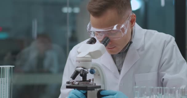 Мужчина-микробиолог изучает образец в лаборатории, изучает микроскоп, специалист по анализу ДНК — стоковое видео