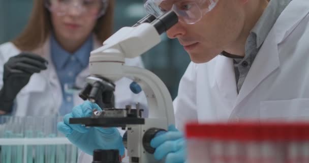 Dois técnicos estão trabalhando em laboratório científico, o homem está pesquisando amostra por microscópio e a mulher trabalhando com produtos químicos. — Vídeo de Stock