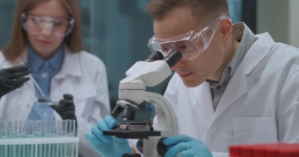 Δύο επιστήμονες εργάζονται στο εγκληματολογικό εργαστήριο, ο άντρας βλέπει στοιχεία στο μικροσκόπιο, η γυναίκα δοκιμάζει υγρά σε δοκιμαστικούς σωλήνες — Αρχείο Βίντεο