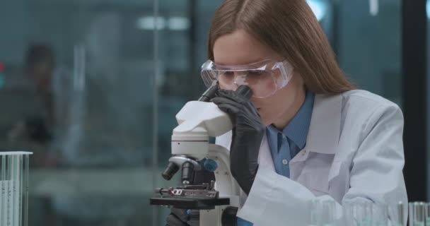 Молодая женщина рассматривает анализ в микроскопе в лабораторных, медицинских и вирусологических исследованиях коронавируса — стоковое видео
