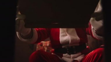Noel Baba kostümü giymiş yaşlı adam, Noel akşamı dekore edilmiş odada, kutudan manzaralı hediye kutusunu açıyor.