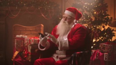 Yaşlı adam Noel Baba kostümü giymiş ve şapka internette akıllı telefondan sörf yapıyor, modern aletler kullanıyor, Xmas akşamı