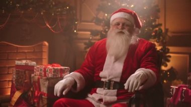 Rahat Noel Baba oturma odasında Noel süsleri ve ağaçla oturmuş kameraya bakıyor. 