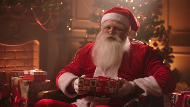 Papá Noel está presentando regalo de Navidad, caja móvil a la cámara y sonriendo alegremente, sentado en la habitación decorada de la casa — Vídeo de stock