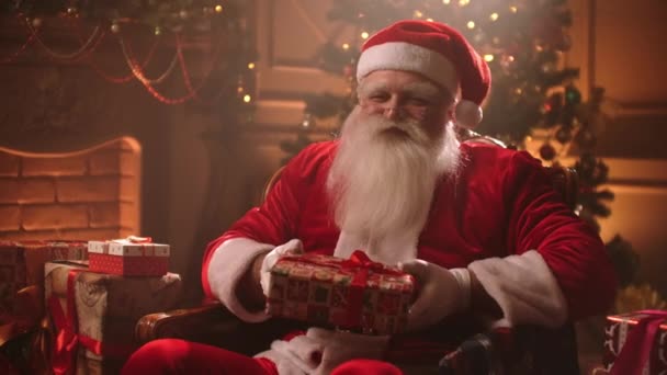 Anciano vestido como Santa Claus está dando regalo de Navidad a la cámara en la sala de estar decorada — Vídeos de Stock