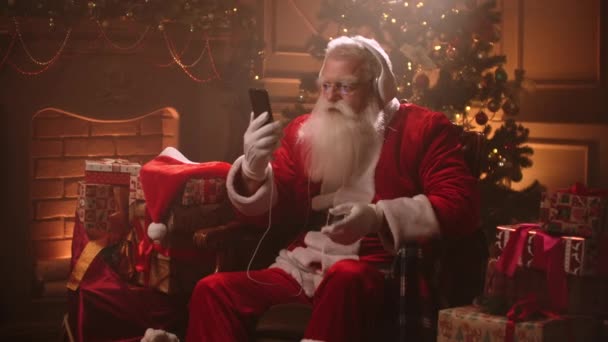 幸せなクリスマスの休日のサンタクロース、古いウィザードは家の中に座っているとヘッドフォンで歌を聴く — ストック動画