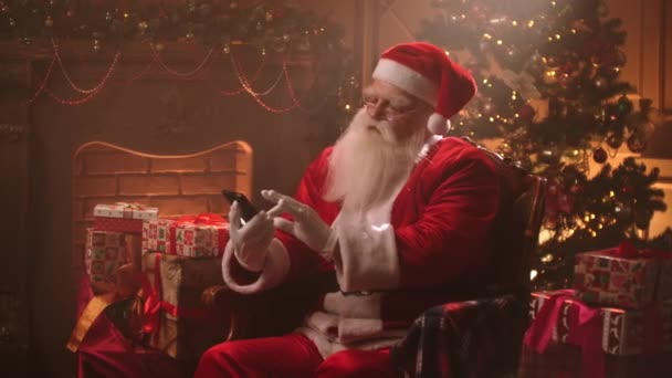Starzec ubrany garnitur Santa Claus i kapelusz jest surfing internet przez smartfona, za pomocą nowoczesnego gadżetu, Boże Narodzenie wieczór — Wideo stockowe