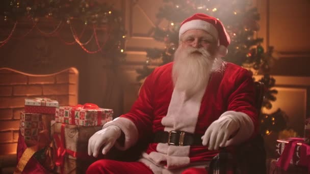 放松的圣诞老人坐在客厅里，手里拿着圣诞装饰品和圣诞树，看着镜头 — 图库视频影像