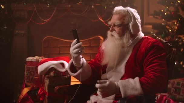 Joyful Santa Claus is listening to music by headphones, looking at smartphone, dancing sitting on chair — Stock Video