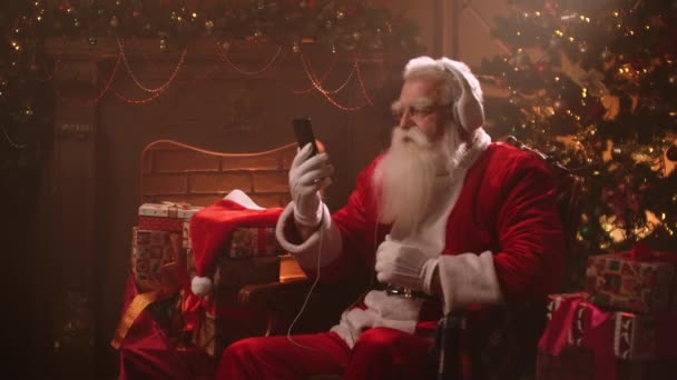 Χαρούμενος Άγιος Βασίλης κάθεται σε διακοσμημένο δωμάτιο το βράδυ των Χριστουγέννων, ακούγοντας μουσική από ακουστικά και smartphone — Αρχείο Βίντεο