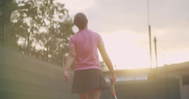 Vista trasera de una jugadora de tenis caminando con una raqueta al atardecer en un mapa en cámara lenta — Vídeos de Stock