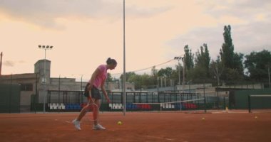 Tenis servisi yapan bir kadının yavaş hareketi. Yavaş çekim: tenis kortunda zıplayan kadın topu bölümü. Sporcu tenis topu servis eder. Genç bir kadın gün batımında tenis raketiyle topa vuruyor.