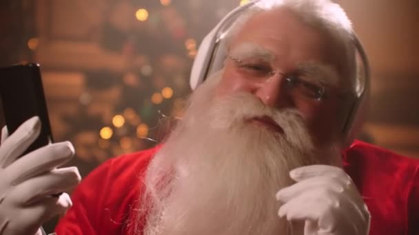 サンタクロースのイメージの俳優は、ワイヤレスヘッドフォンやスマートフォンで音楽を聴いています面白い顔のクローズアップ — ストック動画
