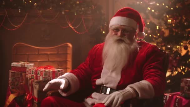 Kind funny Santa Claus is sitting alone at room with Christmas decorations at New year Eve, showing silence sign by finger and mouth — Stock Video