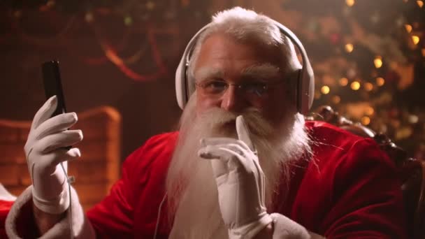 Papai Noel moderno está ouvindo música de Natal por fones de ouvido e smartphone, retrato de assistente — Vídeo de Stock