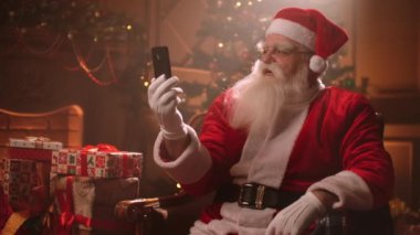 Kırmızı Noel Baba kostümü giymiş bir adam oturma odasında oturmuş akıllı telefonuyla görüntülü sohbet ediyor.