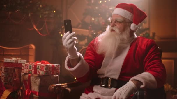 Uomo vestito in abito rosso di Babbo Natale è seduto in soggiorno e chatta con smartphone con video chat — Video Stock