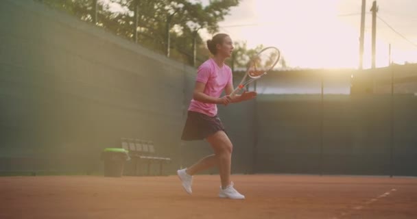 Gün batımında tenis oynayan bir kadın tenis kortunda uçan bir topa vuruyor. — Stok video