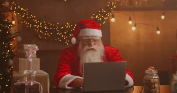 Настоящий Санта Клаус использует новую технологию для общения с детьми, получения почты или списка желаний. Весело работать на ноутбуке и улыбаться, сидя на стуле с камином и рождественской елкой — стоковое видео