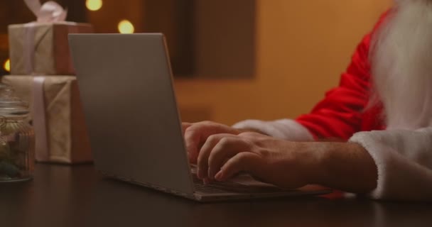 Mains Santa tapant sur le clavier sans fil par table décorée en bois Nouvel An, le Père Noël travaille avec un ordinateur portable regardant par la poste et répondant aux messages aux enfants. — Video