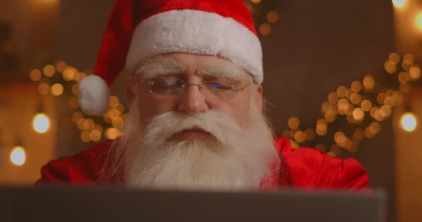 Prawdziwy Święty Mikołaj wykorzystujący nową technologię do komunikacji z dziećmi, odbierania poczty lub listy życzeń. Wesoła praca na laptopie i uśmiech siedząc na krześle z kominkiem i choinką — Wideo stockowe
