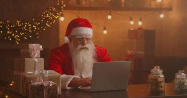 Noel Baba oturma odasında dizüstü bilgisayar kullanıyor. Düşünceli Noel Baba 'nın portresi dekore edilmiş evde modern bir dizüstü bilgisayarla daktilo ediyor.